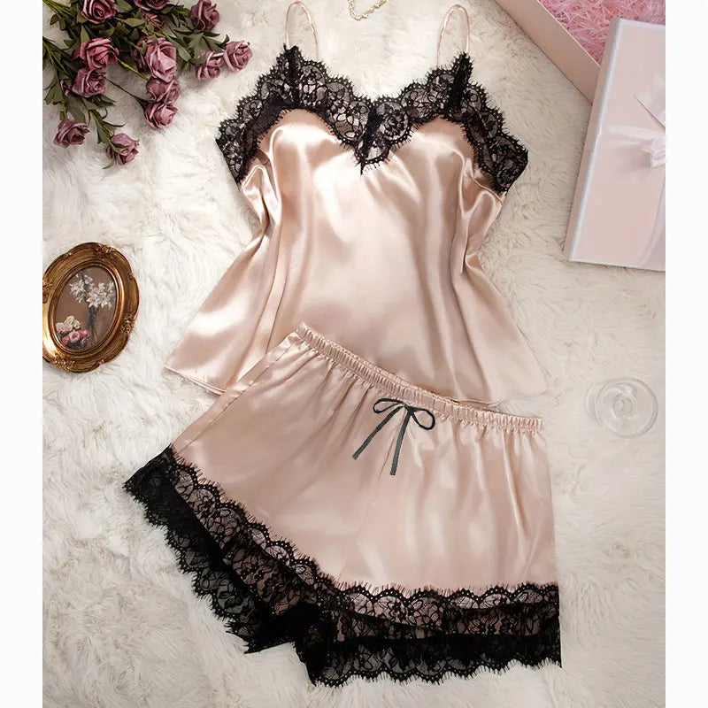 Beige champagne-tone cami and shorts pyjama set laid flat, with matching light straps, black floral lace and a black waist bow